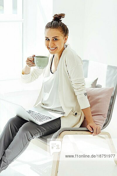 Barfüßige junge Frau sitzt auf einem Stuhl und entspannt sich mit einer Tasse Kaffee  nachdem sie einen Laptop in einem hellen Raum zu Hause benutzt hat
