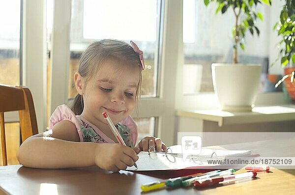Adorable lächelnd kleines Mädchen sitzt am Tisch und Zeichnung mit multicolor Gel Stifte in Notebook  glücklich Kind genießen Kunst Aktivität während der Freizeit zu Hause. Preschooler kreatives Lernen und Entwicklung