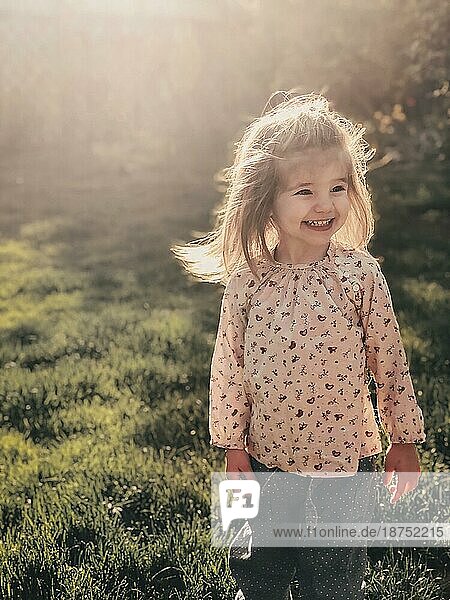 Close up Foto von lächelnden niedlichen kleinen Mädchen von 3 4 Jahren alt mit blonden flauschigen Haaren steht im Freien in hellen warmen Sonnenlicht vor dem Hintergrund der Natur Frühling. Glückliche Kindheit Konzept