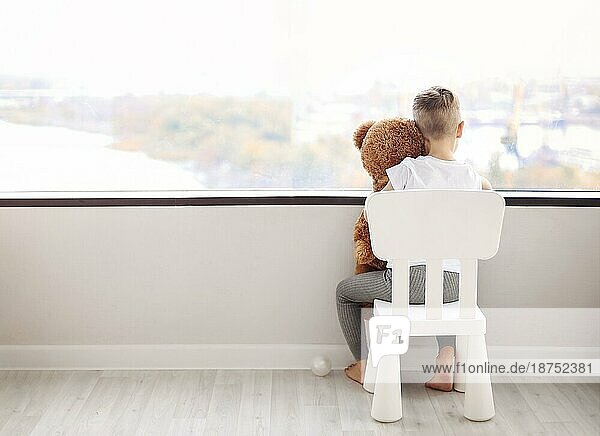 Rückansicht eines kleinen Jungen mit Teddybär  der auf einem Stuhl sitzt und aus dem Fenster auf eine herbstliche Stadtlandschaft schaut  während er zu Hause auf seine Mutter wartet. Kindheit Konzept