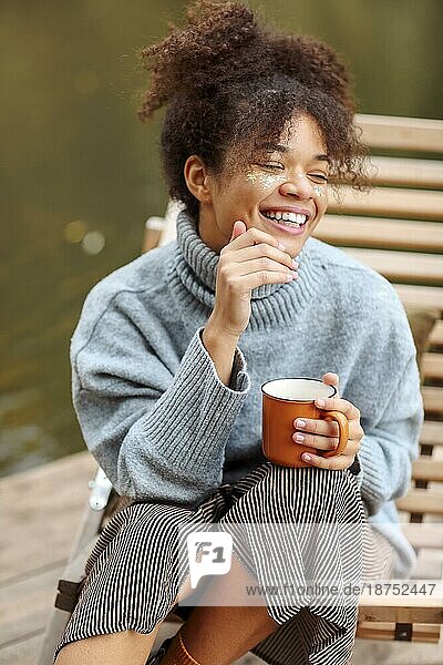 Junge glückliche sorglose afroamerikanische Frau mit einer Tasse heißem Tee  die sich in der Nähe eines Teiches im Herbst entspannt  gemischtrassige Frau im warmen Pullover  die das Herbstwochenende in der Natur genießt. Erholung Konzept