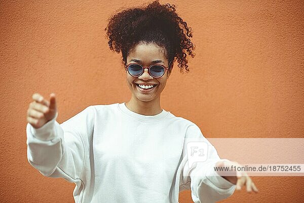 Junge Frau afrikanischer Abstammung mit stilvoller Sonnenbrille  mit lockigem Haar  das zu einem hohen Pferdeschwanz gebunden ist  schaut weg  während sie breit lächelt und gerade  perfekte Zähne zeigt  und posiert vor einem orangefarbenen Wandhintergrund