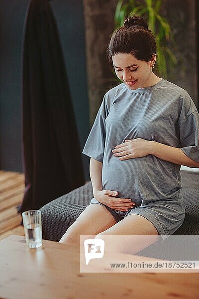Gesunde Gewohnheiten während der Schwangerschaft. Junge glückliche schwangere Frau in Homewear berührt sanft den Bauch  während sie am Tisch mit einem Glas reinen Wassers sitzt und Wasser trinkt  um ihren Körper und ihr Baby gesund zu halten