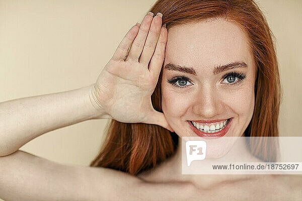 Junge rothaarige Frau mit sauberer Haut  die ihr Gesicht berührt und lächelnd in die Kamera schaut  vor beigem Hintergrund