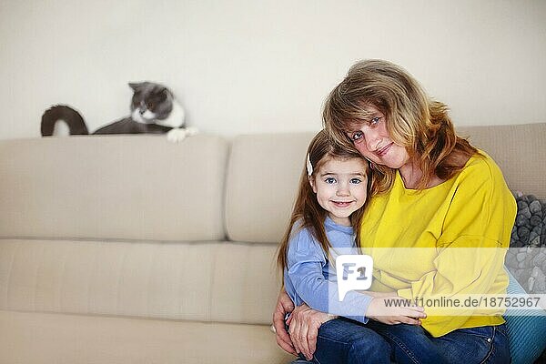Lächelnde Großmutter mittleren Alters und kleine Enkelin  die sich umarmen und in die Kamera schauen  während sie auf dem Sofa sitzen  mit einer niedlichen Katze im Hintergrund  während sie gemeinsam Zeit zu Hause verbringen