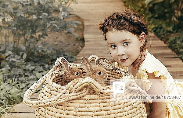 Kleines hübsches Mädchen im Sommerkleid  das draußen auf einem Holzweg vor einem Strohkorb mit Kaninchen sitzt  süßes Kind  das die Kaninchen mit einem leichten Lächeln betrachtet und Zeit mit ihren Lieblingstieren genießt