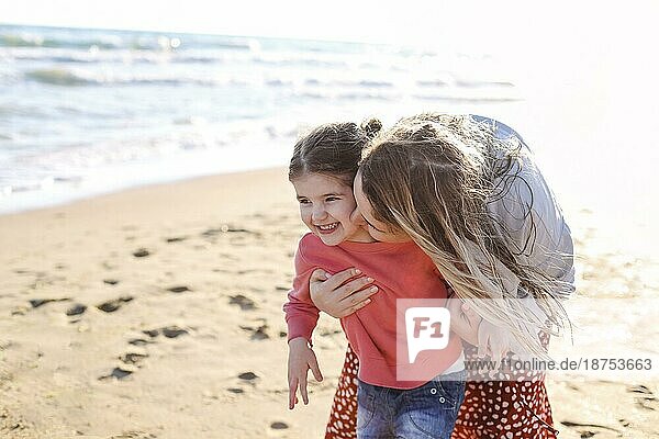 Junge liebende Mutter mit lächelnder Tochter  die ihr am sonnigen Strand entgegenläuft  glückliche Mutter umarmt kleines Mädchen und genießt die gemeinsame Zeit am Sommerstrand. Rückansicht
