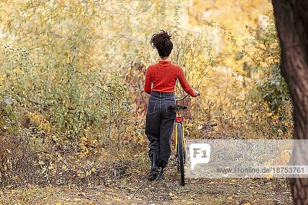 Wochenendaktivität in der Herbstzeit. Junge glückliche lächelnde gemischtrassige Frau mit Fahrrad im Herbst Wald. Rückenansicht der positiven aktiven afrikanischen amerikanischen Frau in Freizeitkleidung Reiten Stadt Fahrrad in der Natur
