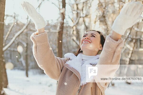 Optimistische junge Frau in modischem Mantel  die lächelnd nach oben schaut und Schneeflocken auffängt  während sie sich am Wochenende auf der Straße amüsiert