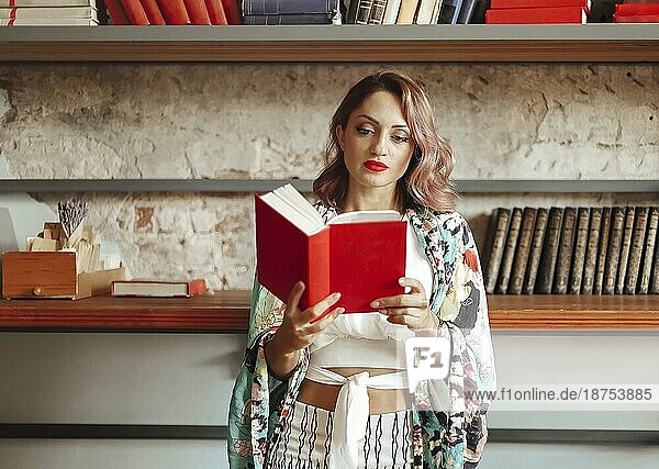 Attraktive selbstbewusste junge Frau in Boho Kleidung hält das Buch  während gegen Bücherregal in der Bibliothek stehen