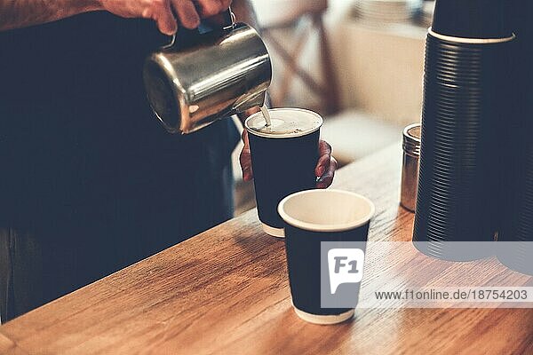 Crop Barista gießt heiße Milch aus Eisen Becher in Kaffeetassen. Hoher Winkel von anonymen professionellen machen Cappuccino und Latte zum Mitnehmen