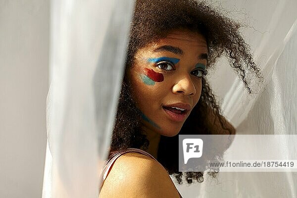 Schöne glückliche afrikanische amerikanische Ethnizität Frau mit kreativen Kunst Make up zur Seite schauen mit angenehmen Lächeln  fröhlich schwarz lockiges Mädchen mit Farbe Abstriche auf Gesicht posiert gegen graue Studio Hintergrund