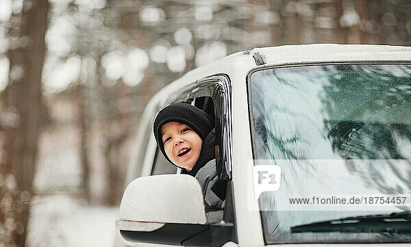 Aufgeregter kleiner Junge mit schwarzer Mütze und warmer Kleidung  der den Kopf aus dem Autofenster streckt und sich umschaut  während er durch den verschneiten Wald in der Wintersaison fährt. Glückliche Kindheit