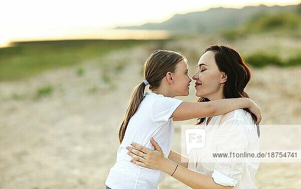 Liebevolle Mutter küsst liebenswertes Mädchen auf die Stirn  während sie im Sommer bei Sonnenuntergang zusammen am Ufer sitzen