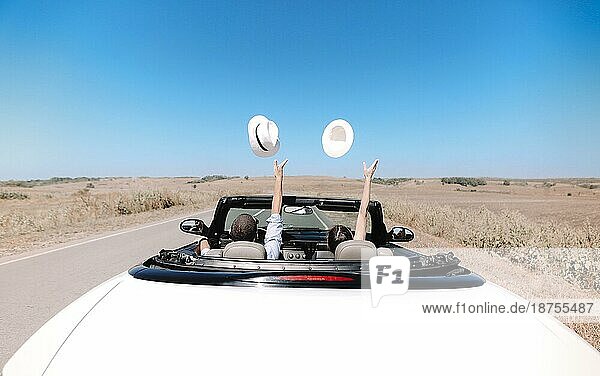 Rückenansicht eines nicht erkennbaren Paares mit Hüten  das die Freiheit genießt  während es in einem Cabrio durch eine grasbewachsene Wüstenlandschaft an einem Sommertag fährt