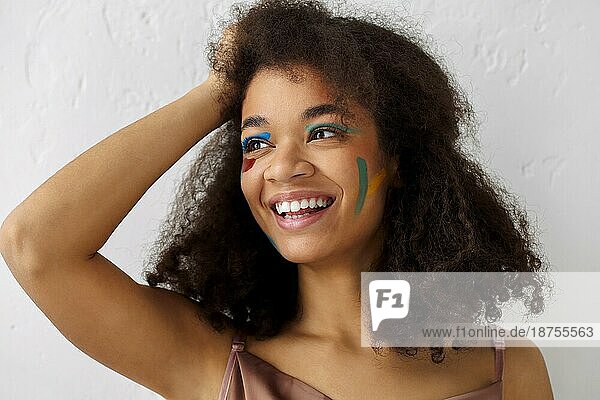 Schöne glückliche afrikanische amerikanische Ethnizität Frau mit kreativen Kunst Make up zur Seite schauen mit angenehmen Lächeln  fröhlich schwarz lockiges Mädchen mit Farbe Abstriche auf Gesicht posiert gegen graue Studio Hintergrund