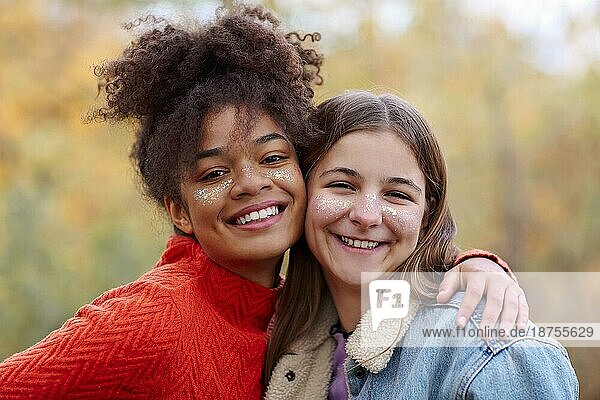 Porträt von zwei glücklichen multirassischen Freundinnen  die in die Kamera lächeln  während sie Zeit in der herbstlichen Natur verbringen  fröhliche Teenagermädchen verschiedener Rassen  die einen gemeinsamen Spaziergang im Herbstwald genießen Weibliche Freundschaft