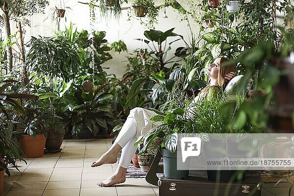 Ruhige Frau mit Händen hinter dem Kopf auf einem Stuhl in einem Raum voller Blumentöpfe mit üppigen Pflanzen liegend
