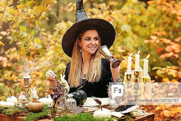 Junge Frau im Hexenkostüm sitzt am Tisch mit magischen Zutaten und macht sich Notizen an einem Herbsttag im Wald