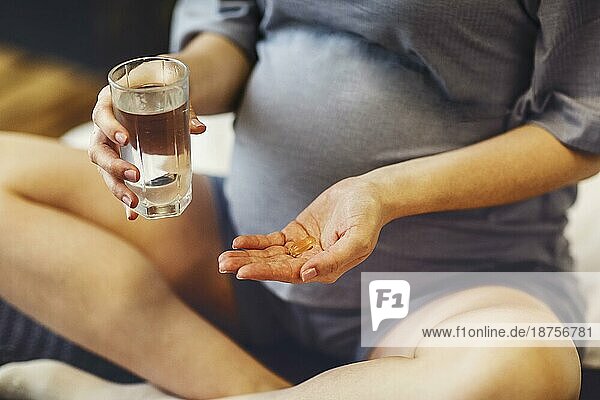 Nahrungsergänzungsmittel während der Schwangerschaft. Junge glückliche schwangere Frau  die pränatale Vitamine einnimmt  mit einem Glas Wasser und einer Pille in der Hand  während sie zu Hause auf dem Bett sitzt. Werdende Mutter beginnt jeden Tag mit Folsäure