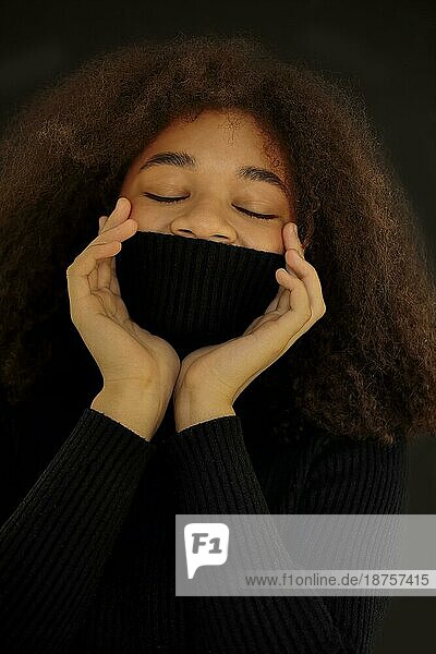 Zufriedene afroamerikanische lockige Frau  die die Augen schließt und das Gesicht mit einem warmen schwarzen Pulloverkragen bedeckt  vordunklem Studiohintergrund  lächelnde Frau  die sich im Rollkragenpullover aufwärmt. Vertikales Foto