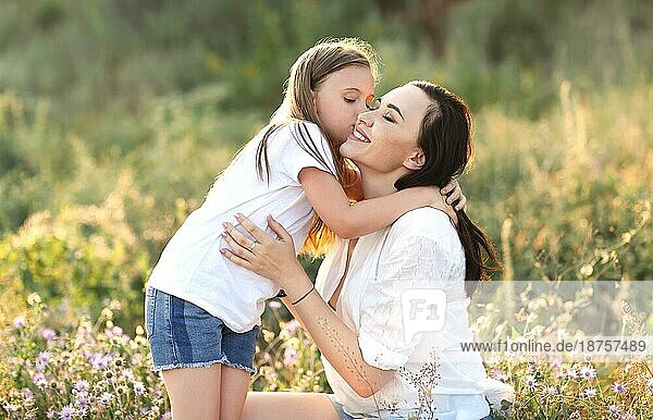Zarte Tochter küsst entzückte Mutter auf die Wange  während sie sich auf einer blühenden Sommerwiese ausruht