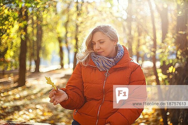 Zufriedene Frau in Oberbekleidung lächelt und betrachtet ein trockenes Blatt an einem sonnigen Wochenendtag im Herbstpark