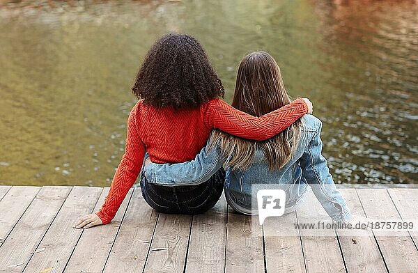 Rückenansicht von zwei glücklichen multirassischen Freundinnen sitzen in der Nähe von Fluss mit Glück  zwei beste weibliche Freunde der verschiedenen Rassen entspannen in der Nähe von Teich im Herbst Wochenende Tag