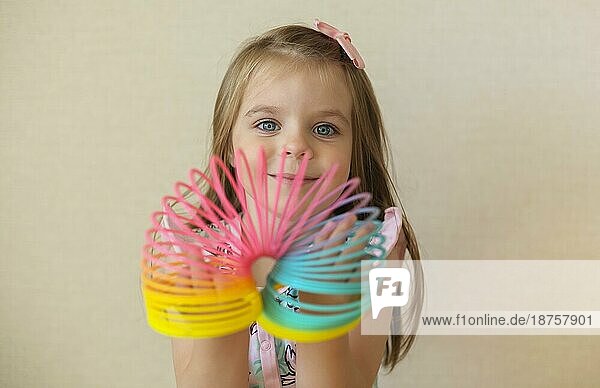 Cute adorable kleines Mädchen spielt mit Kunststoff Regenbogen magische Feder  hält bouncy dehnbar Spielzeug und lächelt in die Kamera. Glückliches Kind vorbeige Hintergrund genießen Freizeit zu Hause