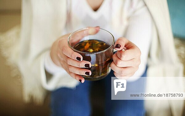 Hohe Winkel der Ernte anonyme weibliche in casual Outfit hält Glas Tasse mit aromatischen Kräutertee beim Entspannen zu Hause