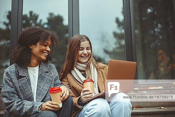 Zwei glückliche  positive  multiethnische Teenagermädchen mit Computer und trinken Kaffee zum Mitnehmen im Freien  schwarzweiße beste Freundinnen  die auf den Laptopbildschirm schauen und lachen  während sie sich im Herbstpark entspannen
