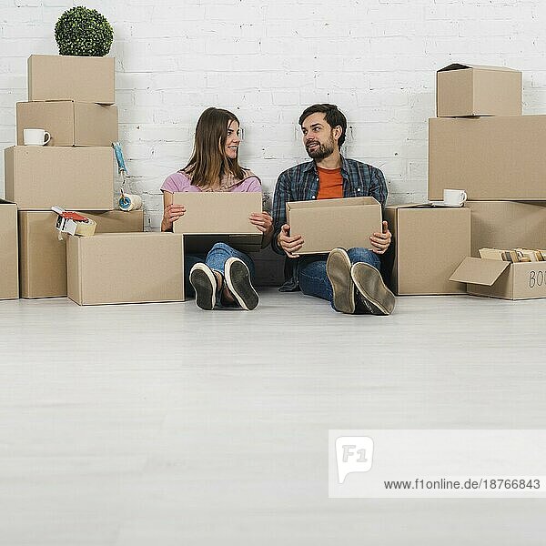 Junges Paar sitzt auf dem Boden und hält Kartons in der Hand und schaut sich gegenseitig an. Foto mit hoher Auflösung