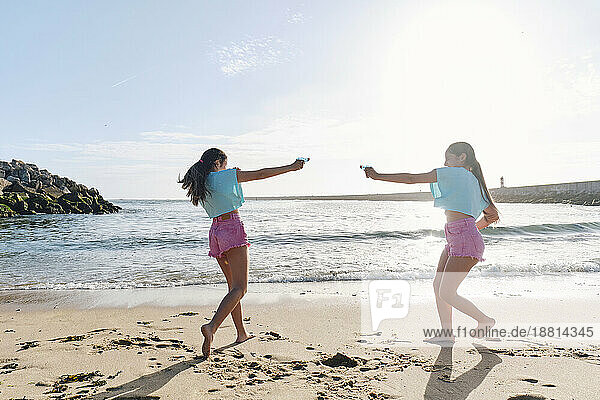 Glückliche Zwillingsschwestern spielen Wasserschlacht mit Spritzpistolen am Strand