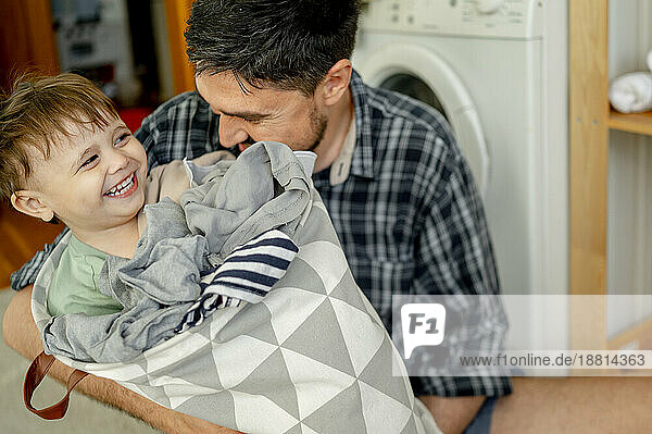 Fröhlicher süßer Junge sitzt im Wäschesack und hat Spaß mit seinem Vater zu Hause