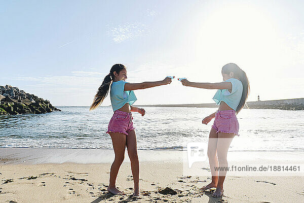 Twin sisters playing with squirt guns on beach at sunny day