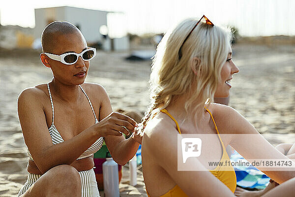 Woman wearing sunglasses braiding hair of friend sitting at beach