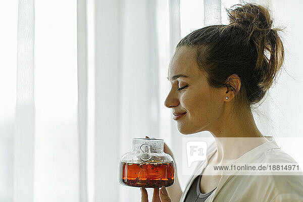 Lächelnde Frau mit geschlossenen Augen  die zu Hause frisch gebrühten Tee riecht