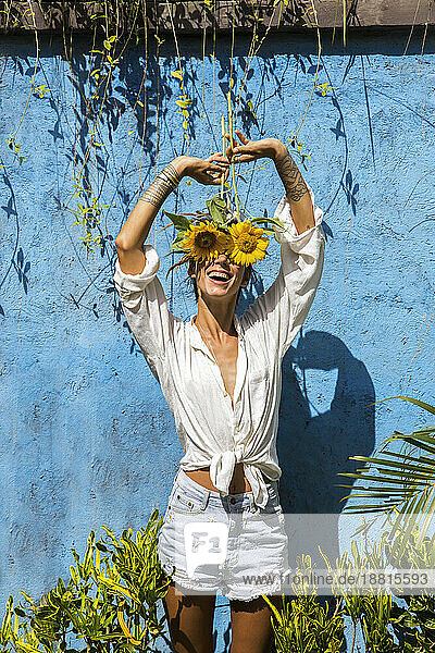 Lächelnde Frau versteckt ihr Gesicht mit Sonnenblumen vor der Wand