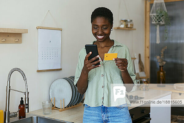 Happy young woman making payment through credit card on smart phone at home