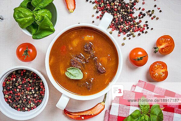 Leckere Tomatensuppe mit Fleisch auf einem weißen rustikalen Holztisch mit frischen Kirschtomaten  Basilikumblättern und trockenem Pfeffer. Zutaten für die Suppe. Ansicht von oben