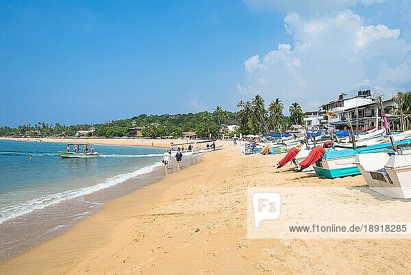 Am Strand eines der wichtigsten Touristenzentren im Südwesten Sri Lankas. Touristen nehmen ein Sonnenbad und treiben Wassersport. Auslegerboote und traditionelle Fischerboote am Strand und vor Anker