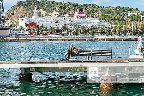 Mann sonnt sich auf einer Bank auf der Rambla Del Mar in Barcelona. Im Hintergrund sind das Hafenbecken mit der Fähre nach Mallorca und der Montjuic zu sehen. Die Rambla del Mar ist eines der Wahrzeichen von Barcelona