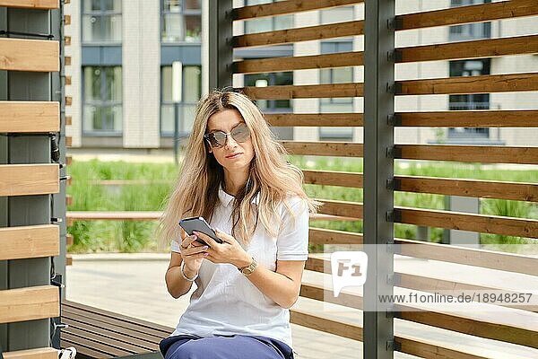 Frau in weißem Hemd und Sonnenbrille auf Bank mit Smartphone