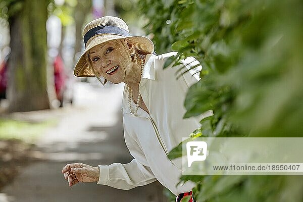 Fröhliche  lächelnde  ältere  sommerlich gekleidete Frau mit Strohhut und Perlenkette  Porträt  Köln  Nordrhein-Westfalen  Deutschland  Europa