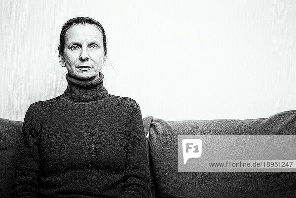 Dramatisches Porträt einer traurigen und deprimierten erwachsenen Frau  die auf einem Sofa sitzt und mit glanzlosen Augen ins Leere schaut. Negativer Raum für Text
