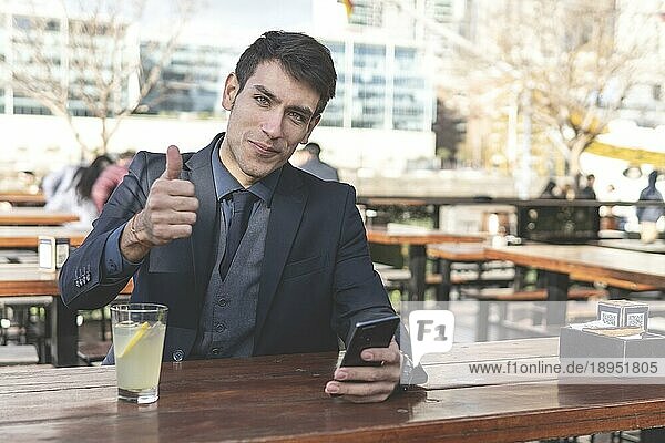 Geschäftsmann sitzt an einer Bar im Freien und trinkt Limonade  während er in die Kamera schaut und einen Daumen nach oben zeigt
