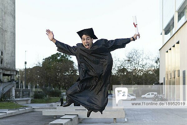 Ein Mann  der vor kurzem seinen Abschluss gemacht hat  in Kappe und Talar  zeigt springend seinen Abschluss und feiert
