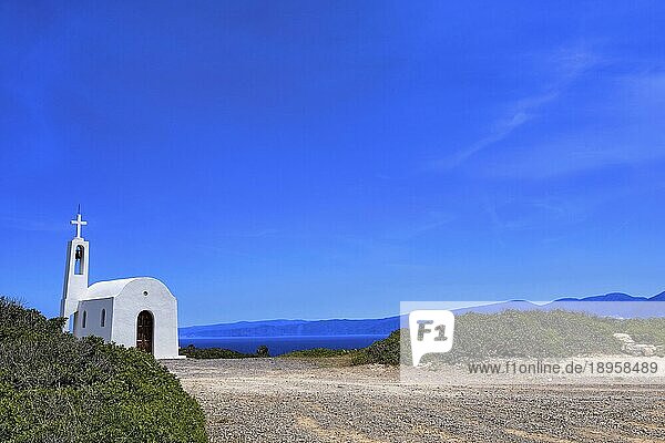 Weiße griechisch orthodoxe Kapelle oder Kirche auf einem Hügel am Meer und die Straße dorthin gegen den klaren blauen Himmel an einem sonnigen Tag. Traditionelles religiöses Gebäude. Typische griechische Landschaft
