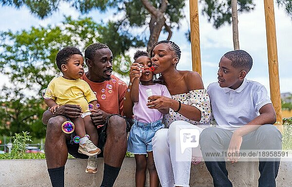 Schwarzafrikanische Familie mit Kindern auf dem Spielplatz  die sich beim Blasen von Seifenblasen neben den Bäumen im Park vergnügen