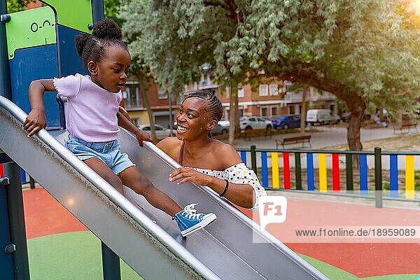 Afrikanische schwarze ethnische Mutter hat Spaß mit ihrer Tochter in der Quietsche des Spielplatzes des Stadtparks in den Sonnenuntergang
